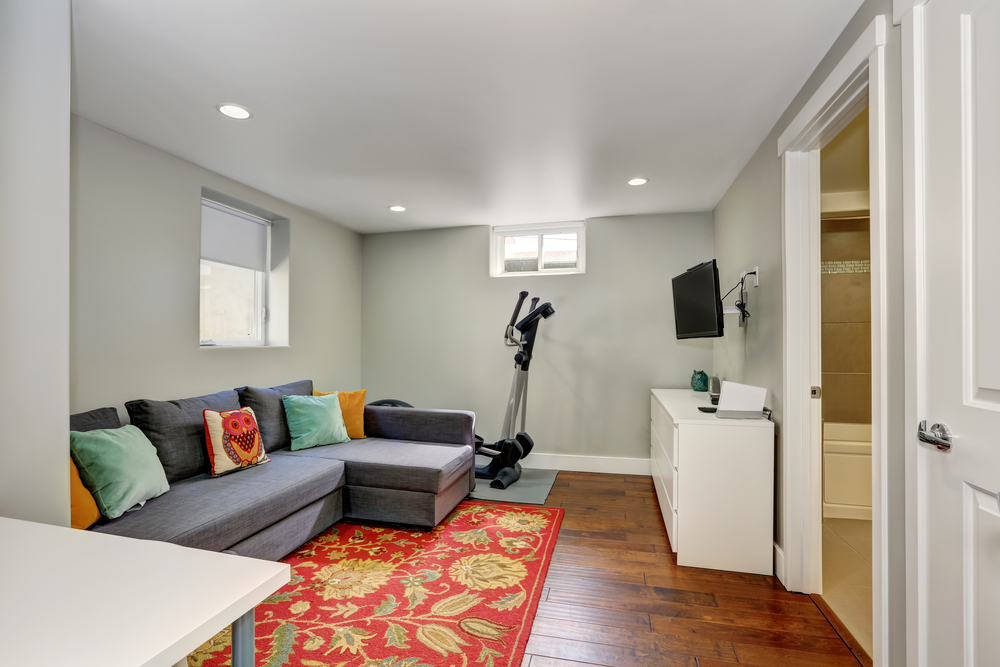 Sitting room interior with sport equipment and TV in the basement. There is a grey sofa with colorful pillows on a red floral accent rug. 